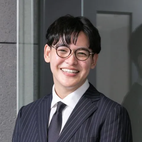 眼鏡をかけた男性が笑顔で写っている写真です。彼はストライプのスーツとネクタイを着用しています。