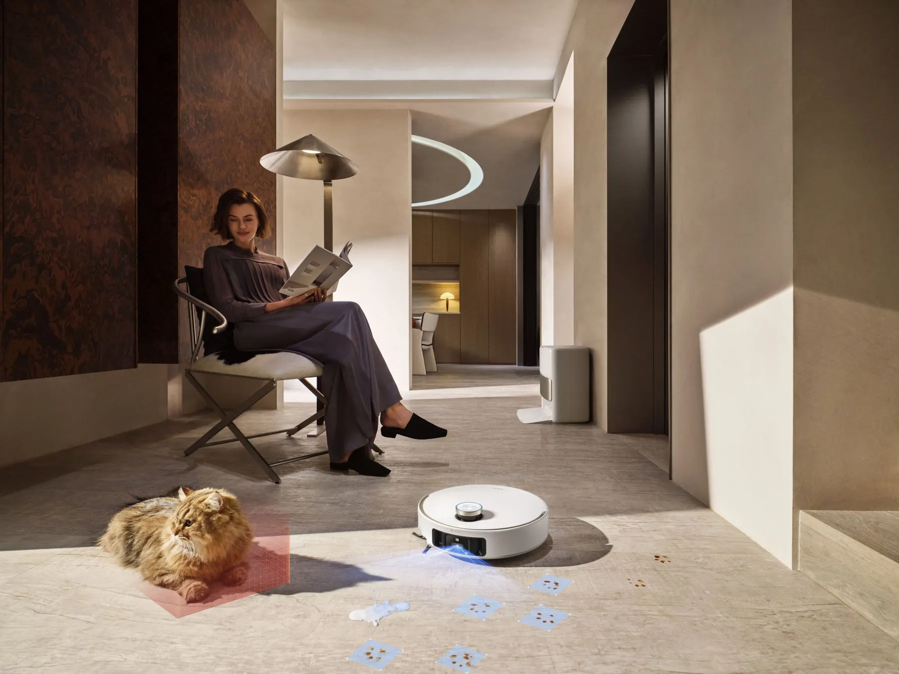 女性が本を読みながら椅子に座り、猫とロボット掃除機が近くにある、モダンなリビングルームのシーン。