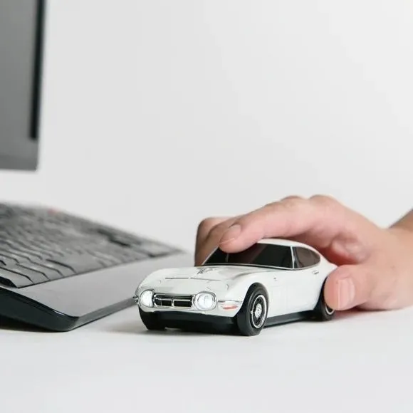 白いスポーツカーの形をしたマウスを操作している手のクローズアップ写真。背景にはキーボードとモニターの一部が見える。