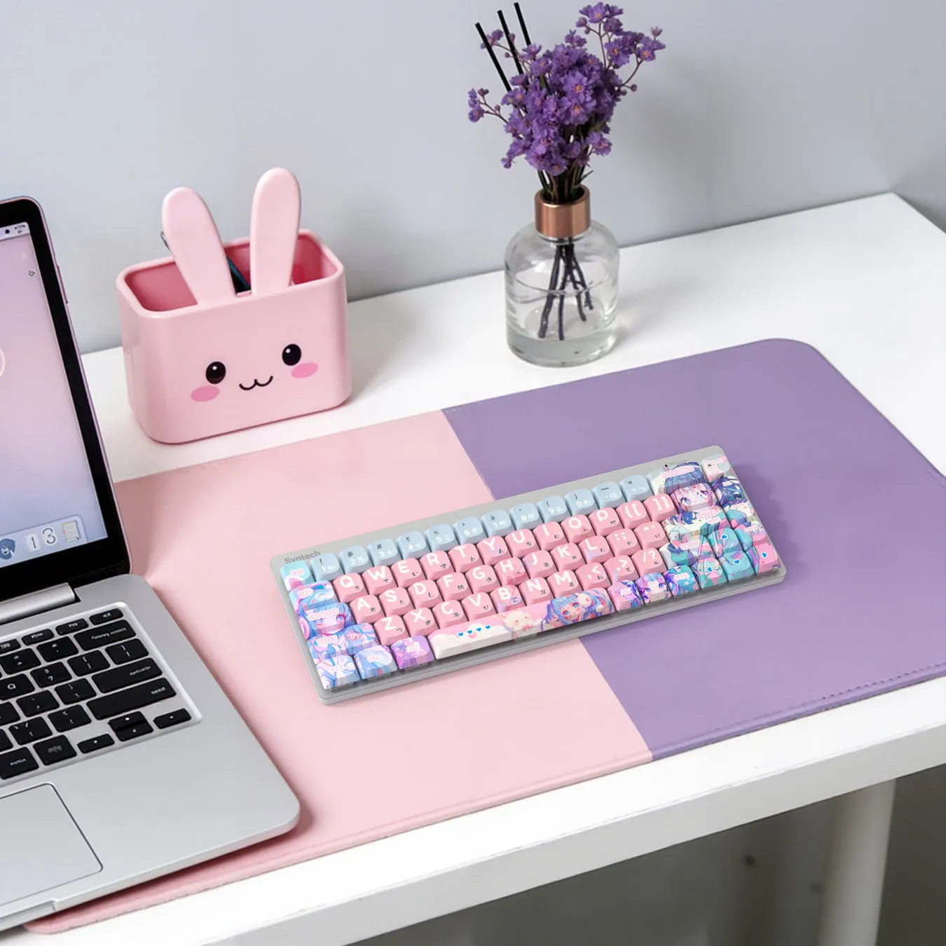 ピンクと紫のデスクマットの上に、アニメ風のキーキャップが付いたメカニカルキーボード、ウサギの形をしたペン立て、花瓶に入った花、ノートパソコンが置かれた、可愛らしい雰囲気のデスク周り。