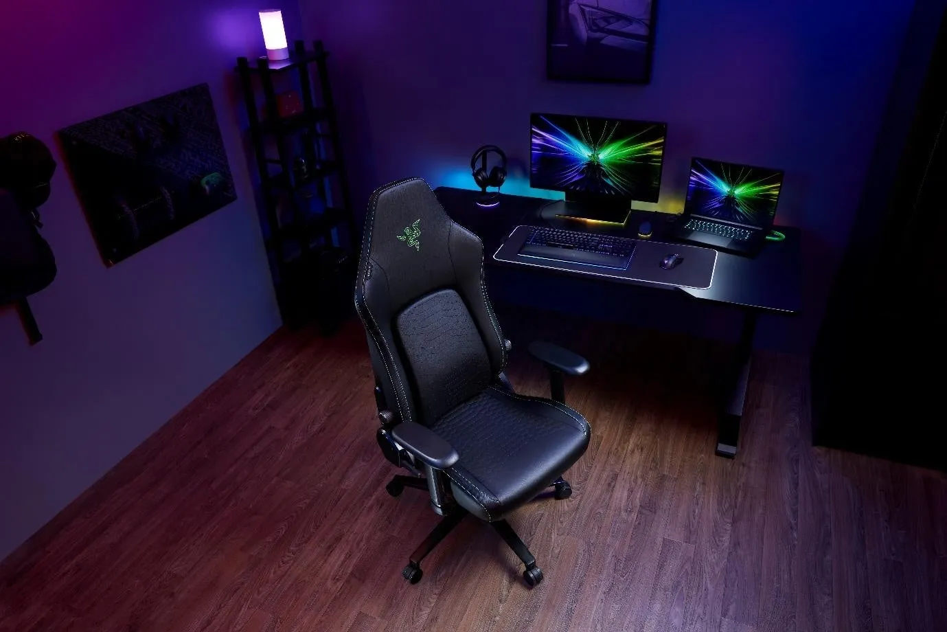 紫色の照明が施されたゲーミングルームにRazerのゲーミングチェアが置かれています。デスクにはモニター、キーボード、マウス、ノートパソコン、ヘッドフォンが配置されています。